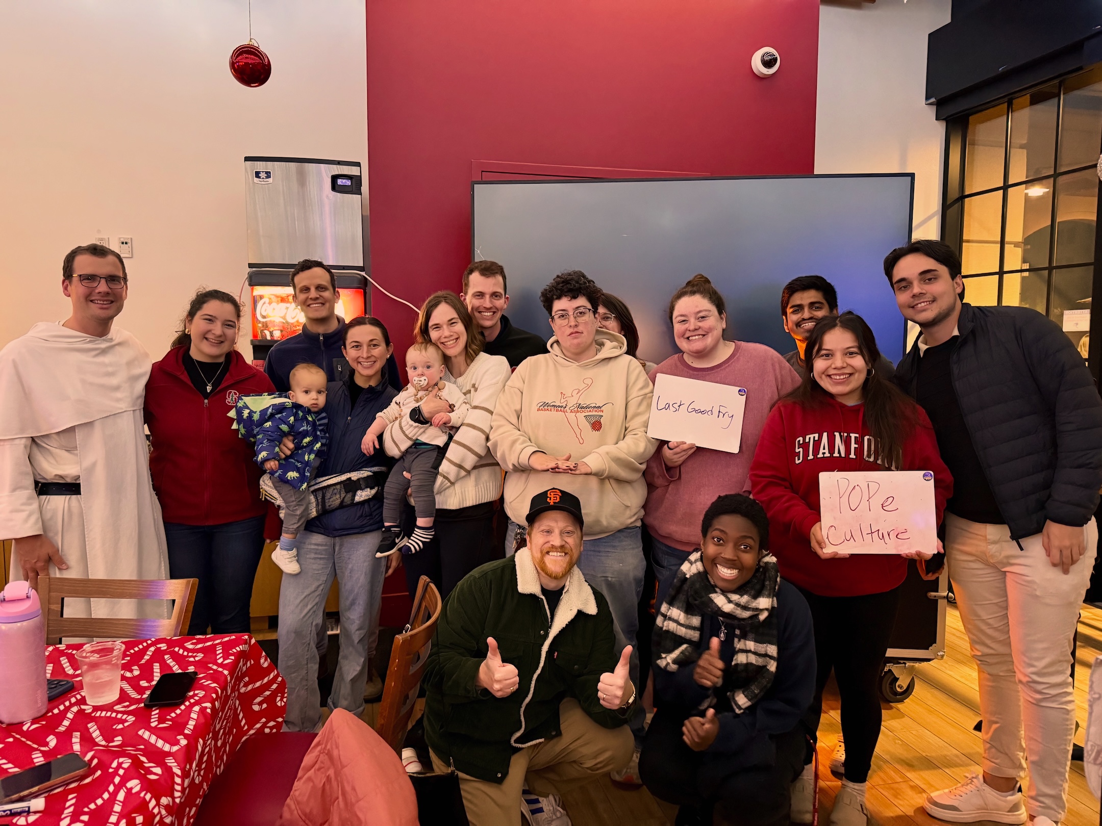 Members of Stanford Universityʼs Catholic communuity gather for grad trivia night. From left to right: Father Chrysostom Mijinke, OP, Isabella McCormick, Andrew Cheshire, Marlena Cheshire holding Michael Cheshire (baby), Caneel Cooper holding Warren Cooper, Matteus Cooper, Unknown, Maria Moore, Nevin Aresh, Lisa Archuleta, Gabriel. Bottom: Bo Lotti, Uwade Akhere. | Photo courtesy of Lisa Archuleta,